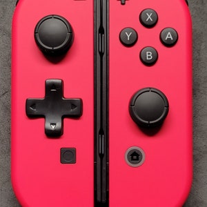 May include: A bright pink Nintendo Switch Joy-Con controller with black buttons and a black d-pad. The controller has a home button, a capture button, and a minus button.