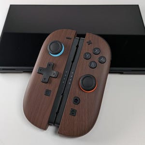 Könnte beinhalten: Ein Controller in brauner Holzmaserung mit blauen und orangen Akzenten, mit einem Steuerkreuz, Tasten und einem schwarzen Bildschirm im Hintergrund. Der Controller ist für Spiele konzipiert.