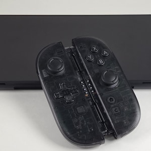 May include: A black video game console with a matching pair of translucent black controllers. The controllers feature a directional pad, two analog sticks, and buttons labeled with X, Y, A, and B, along with home and capture icons on the right side.