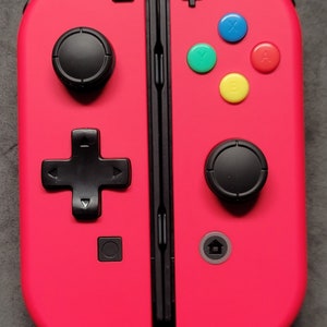 May include: A bright pink Nintendo Switch controller with black buttons and a black d-pad. The controller has a plus sign on the right side and a minus sign on the left side. The controller has four colored buttons: blue, green, yellow, and red.