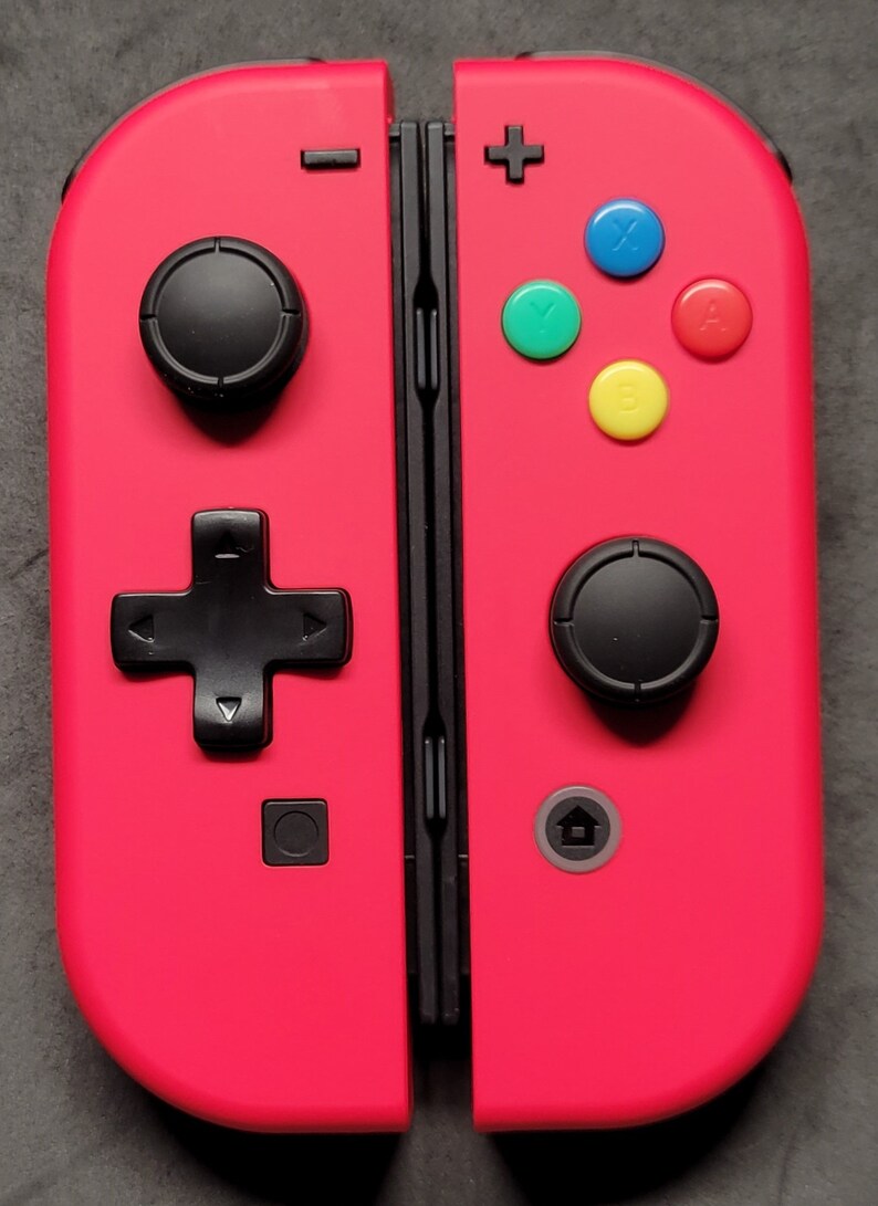 May include: A bright pink Nintendo Switch controller with black buttons and a black d-pad. The controller has a plus sign on the right side and a minus sign on the left side. The controller has four colored buttons: blue, green, yellow, and red.