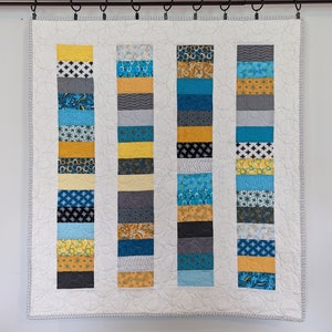 May include: A white quilt with a geometric pattern of blue, yellow, and gray fabric strips. The quilt is hanging on a white wall with a black rod.
