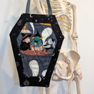 May include: A black coffin-shaped tote bag with a quilted design. The bag features a print of three witches playing cards. The bag has a black handle and is decorated with stars and crescent moons. The background includes a skeleton.