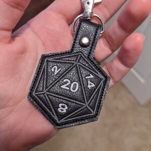 The black vinyl keychain variant shaped like a twenty-sided die with white stitching and numbers 2, 8, 20, and 4 visible. The keychain has a silver metal clasp to clip onto any bag or backpack.