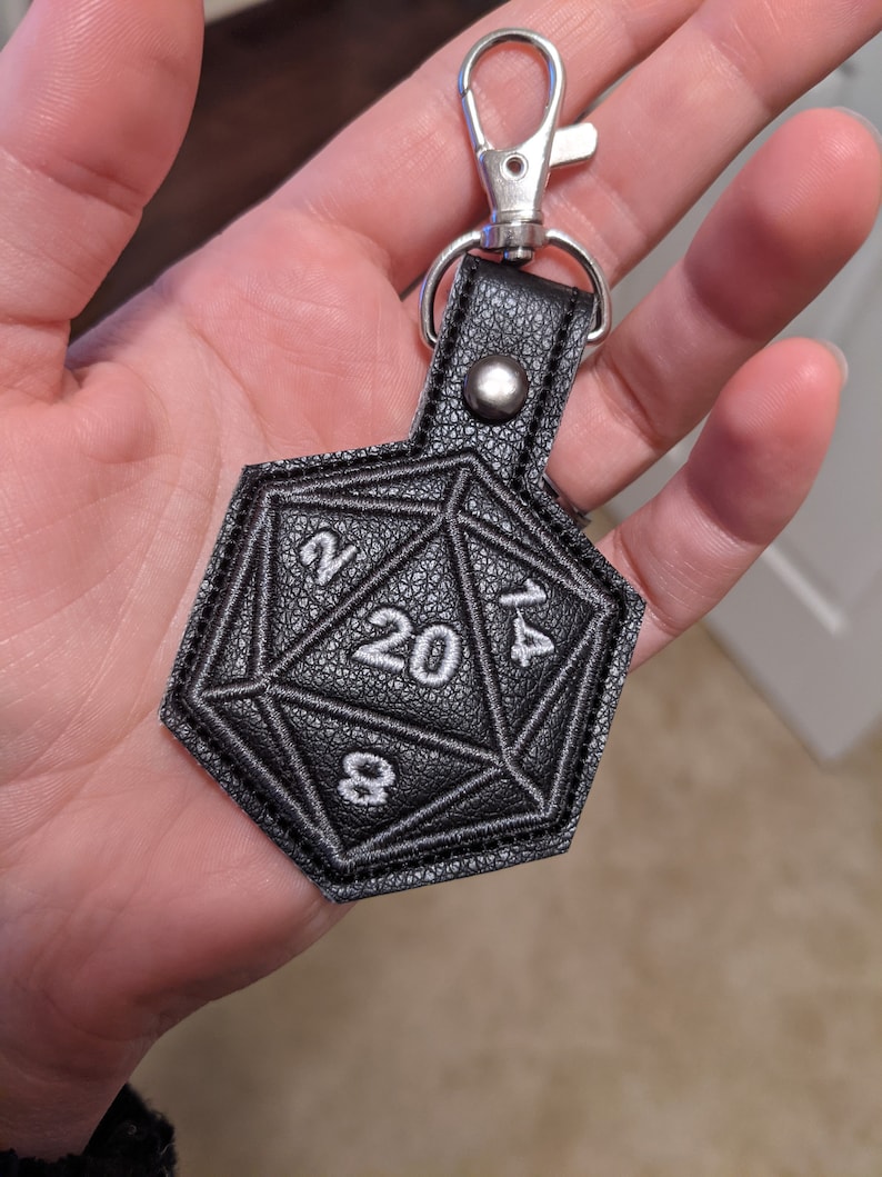 The black vinyl keychain variant shaped like a twenty-sided die with white stitching and numbers 2, 8, 20, and 4 visible. The keychain has a silver metal clasp to clip onto any bag or backpack.