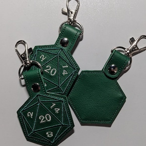 The hunter green vinyl keychains with silver metal clasp. This picture shows both front and backs of the keychain.  The front features embroidered details to look like a twenty-sided die, with a critical roll of 20 being most visible.  The back is plain.  A silver metal clip is used to attach onto bags, purses, backpacks, luggage, and more.