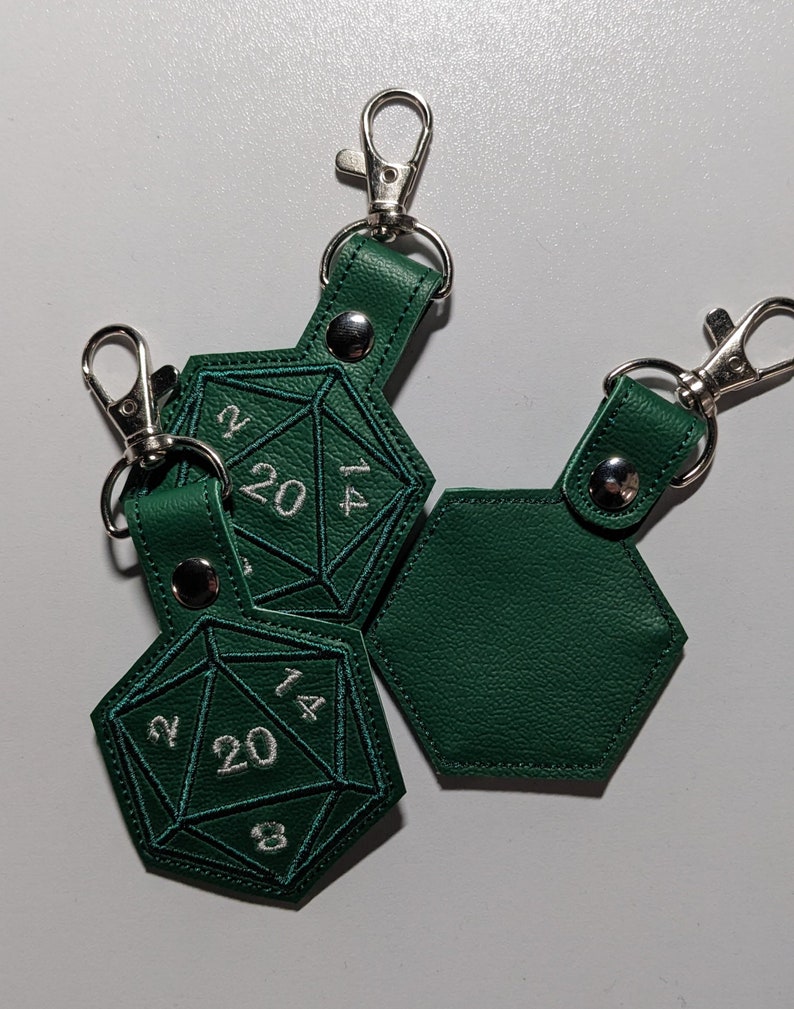 The hunter green vinyl keychains with silver metal clasp. This picture shows both front and backs of the keychain.  The front features embroidered details to look like a twenty-sided die, with a critical roll of 20 being most visible.  The back is plain.  A silver metal clip is used to attach onto bags, purses, backpacks, luggage, and more.