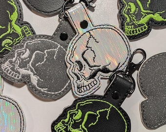 Könnte beinhalten: Eine Sammlung von schädelförmigen Schlüsselanhängern aus schwarzem lederähnlichem Material mit neongrünen Stickereidetails sowie ein Schlüsselanhänger aus reflektierendem, irisierendem Material mit schwarzen Stickkonturen und schwarzen Metallverschlüssen.