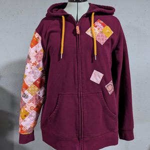 May include: A maroon zip-up hoodie with a yellow zipper and yellow drawstrings. The hoodie has patchwork fabric on the sleeves and a small patchwork fabric square on the front.