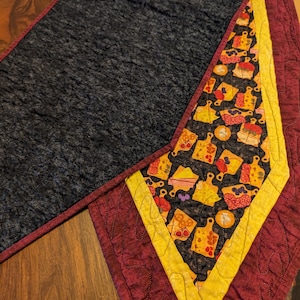 May include: A quilted table runner with a black background and a colorful pattern of pies and pastries. The runner has a burgundy border with a yellow accent.