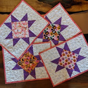 May include: Set of four square quilted placemats. Each features a central star design in purple, with a patterned center square in pink, orange, or black. The edges are trimmed with a pink border, and the background is white.