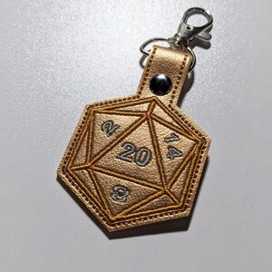 The gold foil vinyl keychain variant with a silver metal clasp. The keychain is shaped like a twenty-sided and embroidered with silver and gold thread.