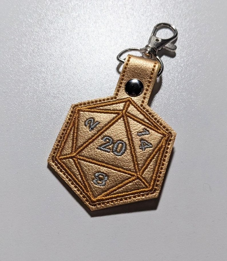 The gold foil vinyl keychain variant with a silver metal clasp. The keychain is shaped like a twenty-sided and embroidered with silver and gold thread.