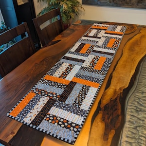 May include: A Halloween-themed table runner with a patchwork design featuring black, white, orange, and gray fabric squares. The runner is laid on a wooden table.