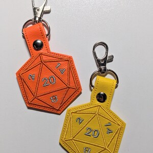 Two vinyl keychains shaped like a twenty-sided die, one is orange and the other is yellow. Both keychains have silver metal clasps. The numbers 2, 4, 8, 14, and 20 are embroidered on the dice in silver thread.