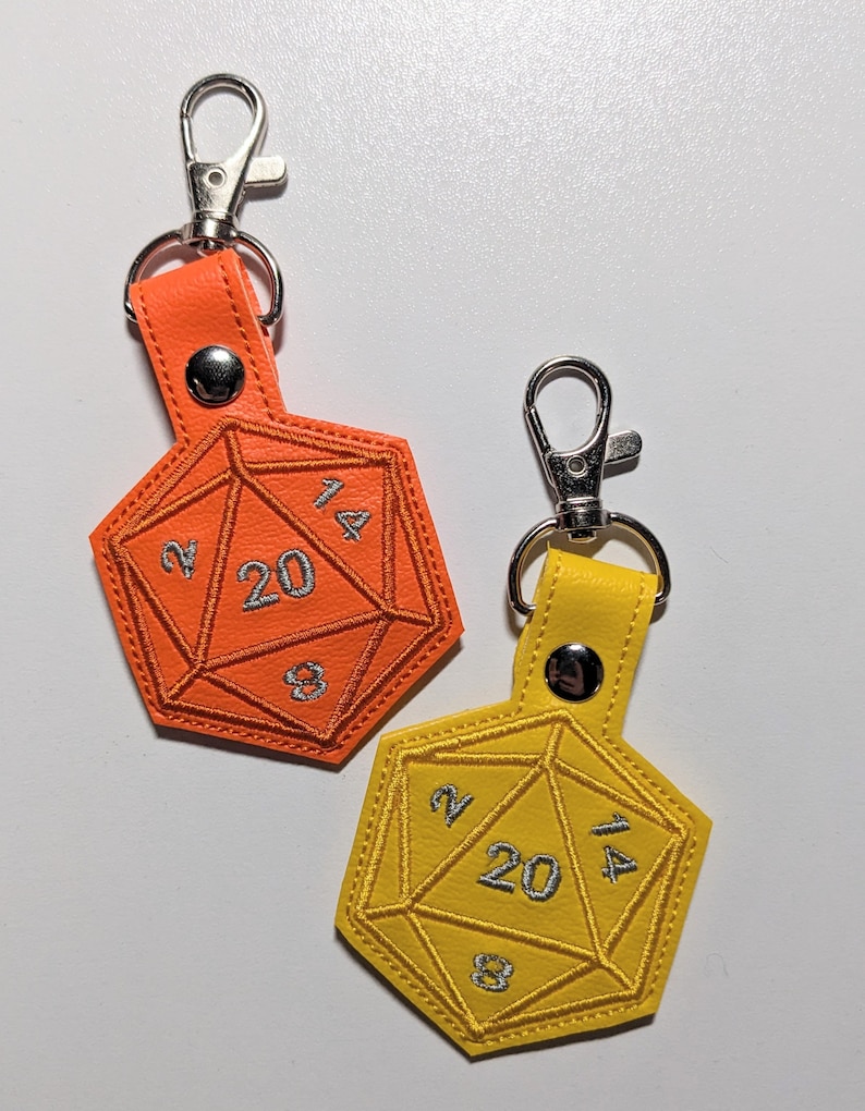 Two vinyl keychains shaped like a twenty-sided die, one is orange and the other is yellow. Both keychains have silver metal clasps. The numbers 2, 4, 8, 14, and 20 are embroidered on the dice in silver thread.
