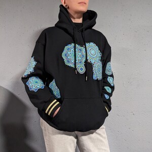 May include: Black hoodie with a large blue and teal mandala design in the shape of Africa on the front. Additional mandala designs are on the sleeves. The hoodie has a kangaroo pocket and gold stripes on the cuffs.