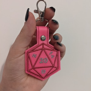 The pink vinyl variant keychain with a silver metal clasp. The keychain is shaped like a twenty-sided die and is embroidered with silver thread for the numbers, and a matching pink thread.
