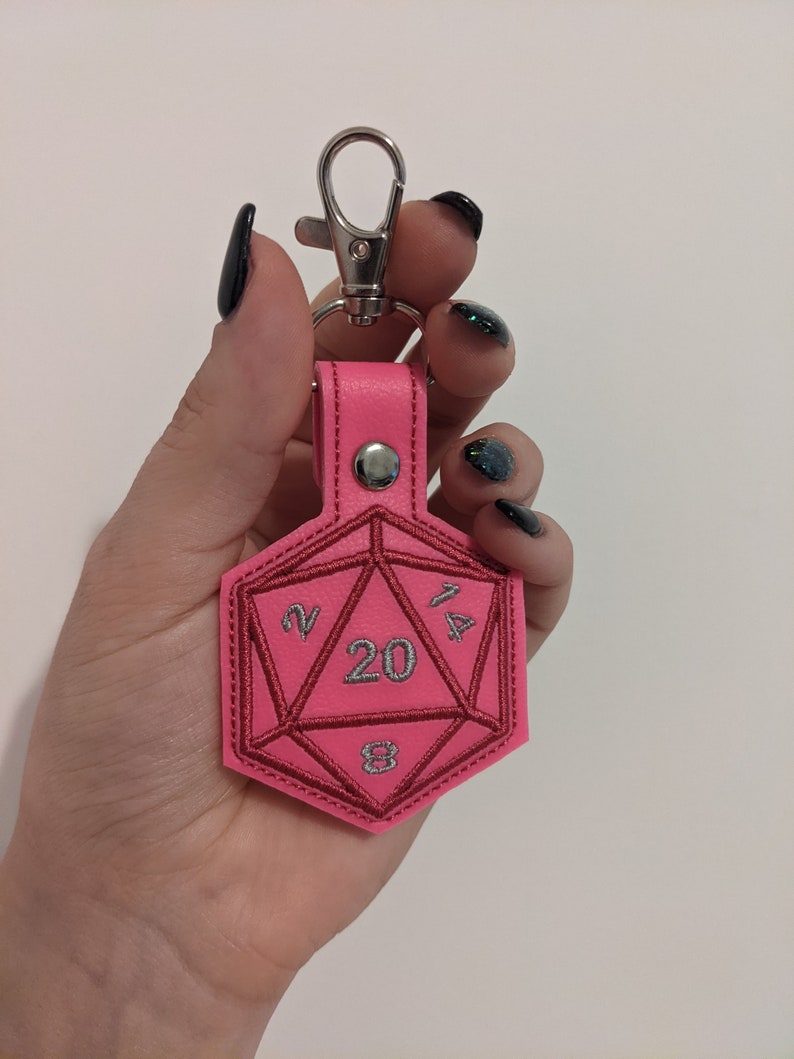 The pink vinyl variant keychain with a silver metal clasp. The keychain is shaped like a twenty-sided die and is embroidered with silver thread for the numbers, and a matching pink thread.