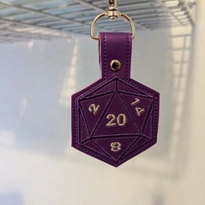 The purple vinyl variant of the D20 embroidered key fob.  The keychain is clipped onto a metal grate for this photograph.