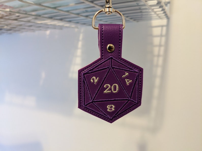 The purple vinyl variant of the D20 embroidered key fob.  The keychain is clipped onto a metal grate for this photograph.