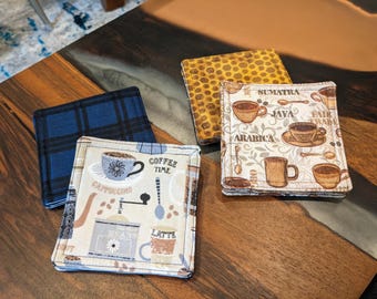Könnte beinhalten: Eine Sammlung quadratischer Stoffuntersetzer mit Kaffeemotiven. Ein Untersetzer hat ein blaues Karomuster, ein anderer einen gelb-braunen Tierdruck, und zwei zeigen Kaffeetassen, Bohnen und Text wie "Cappuccino" und "Coffee Time".