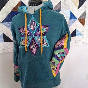May include: Teal blue hoodie with a large colorful star-shaped patch on the front and smaller patches on the sleeves. The star patch features a variety of images including a mermaid, a frog, and a skull. The text "remarkable" is printed on the patch.