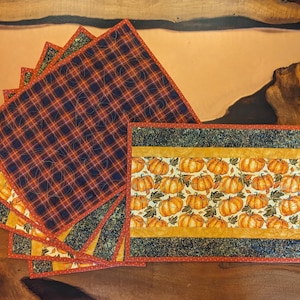 May include: A set of quilted placemats with a fall theme. The placemats feature a plaid pattern in shades of orange, navy, and red, and a pumpkin pattern with orange pumpkins on a cream background. The placemats are rectangular.