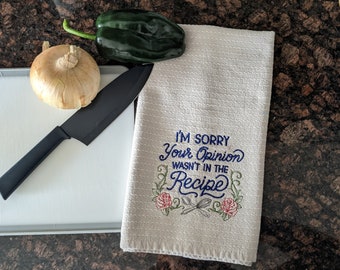 Könnte beinhalten: Ein beiges Küchentuch mit dem gestickten Text "I'M SORRY Your Opinion WASN'T IN THE Recipe" und floralen Akzenten. Ein schwarzes Messer, eine Zwiebel und eine grüne Paprika liegen auf einem Schneidebrett.
