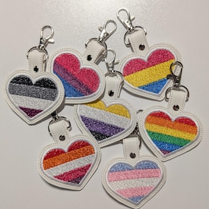 May include: A collection of heart-shaped keychains with various LGBTQ+ pride flag designs. Each keychain features a white leather backing, a silver clasp, and embroidered flags in vibrant colors, including rainbow, bisexual, and transgender flags.