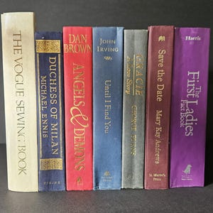 May include: A collection of hardcover books with various colored spines. Titles include "The Vogue Sewing Book", "Duchess of Milan", "Angels & Demons", "Until I Find You", "Gracie", "Save the Date", and "The First Ladies Fact Book".