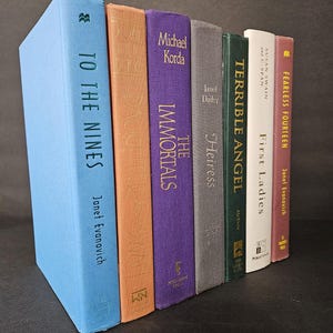 May include: A collection of hardcover books with various colored spines, including blue, orange, purple, gray, green, white, and red. Titles and authors are visible, such as "To The Nines" by Janet Evanovich and "The Immortals" by Michael Korda.