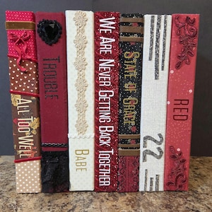 May include: A row of six decorative book spines featuring various fabric textures, lace, and embroidery. The spines display text including All Too Well, Trouble, Babe, We Are Never Getting Back Together, State of Grace, 22, and Red in diverse fonts.