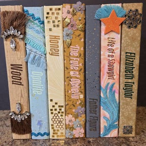 May include: A collection of seven decorative book spines featuring varied textures and embellishments. Titles include Wood, Opalite, Honey, The Fate of Ophelia, Father Figure, Life of a Showgirl, and Elizabeth Taylor, adorned with tassels, gems, and fabric.