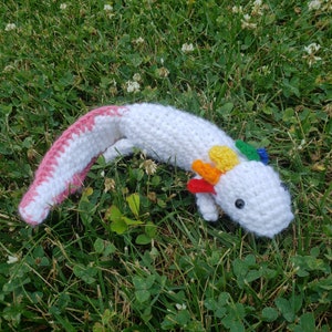May include: A white crocheted axolotl with a pink tail and rainbow colored accents. The axolotl is lying on a bed of green grass.