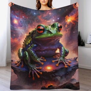 Könnte beinhalten: Eine weiche Decke mit einer Illustration eines Frosches mit roten Augen vor einem farbenfrohen Nebelhintergrund. Der Frosch ist grün und lila, mit orangefarbenen Akzenten. Ein gemütliches Wohnaccessoire.