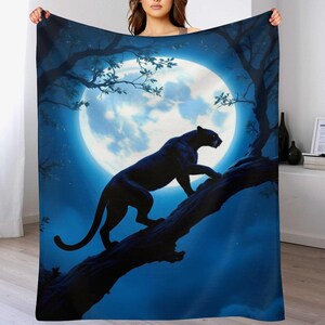 May include: A dark blue blanket featuring a black panther climbing a tree branch against a backdrop of a full moon. The design includes tree branches and leaves, creating a nighttime scene. The blanket is held up by a person.