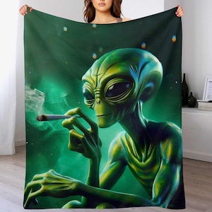 May include: A green blanket with a detailed illustration of a green alien smoking a cigarette. The alien is set against a green and black background with a smoky effect. The blanket is held up by a person.