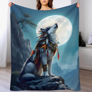 Peut inclure: Un plaid rectangulaire présentant une illustration graphique d'un loup hurlant à la pleine lune. Le loup est orné de plumes colorées et de bandes décoratives, assis sur une falaise rocheuse devant un paysage de montagnes bleues.