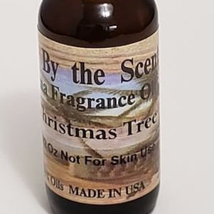 Puede incluir: Una pequeña botella de vidrio marrón con una tapa negra. La etiqueta dice "By the Scents" y "Fragrance Oil Christmas Tree" con una imagen de una escena de bosque nevado. La etiqueta también dice "1 oz Not For Skin Use" y "Oils Made in USA".