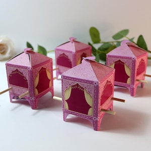 Puede incluir: Cuatro cajas decorativas de papel rosa con dise&ntilde;os ornamentados, que se asemejan a peque&ntilde;os carruajes. Cada caja tiene un interior magenta, detalles dorados y clavijas de madera. Las cajas est&aacute;n dispuestas sobre una superficie blanca.