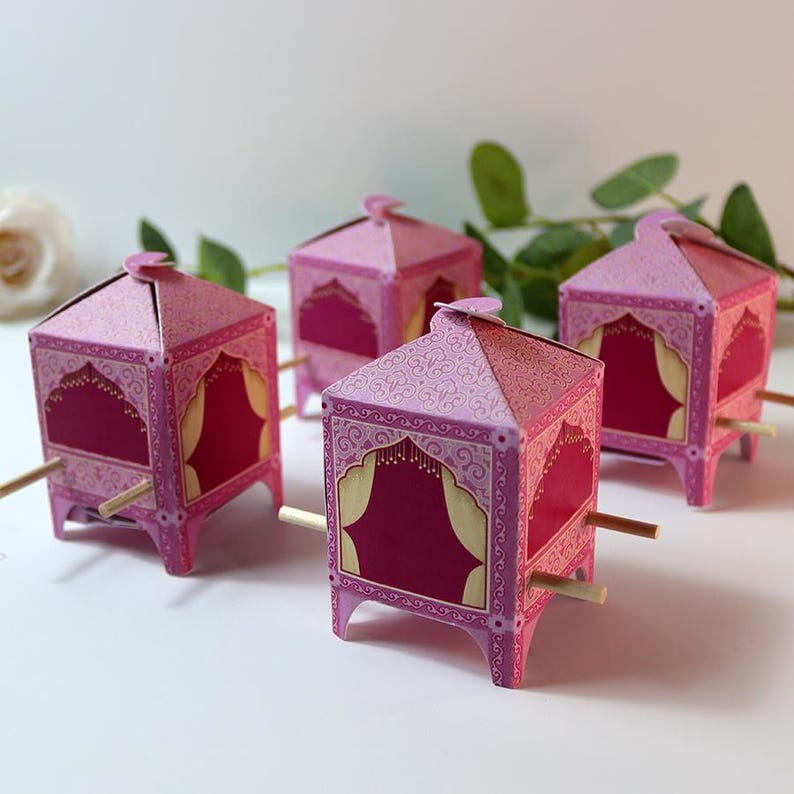 Puede incluir: Cuatro cajas decorativas de papel rosa con dise&ntilde;os ornamentados, que se asemejan a peque&ntilde;os carruajes. Cada caja tiene un interior magenta, detalles dorados y clavijas de madera. Las cajas est&aacute;n dispuestas sobre una superficie blanca.