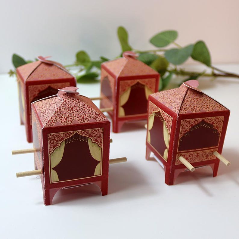 Puede incluir: Cuatro cajas decorativas de papel rojo con forma de palanquines. Cada caja presenta un techo estampado, detalles dorados y burdeos ornamentados y clavijas de madera. Las cajas est&aacute;n dispuestas sobre una superficie blanca con vegetaci&oacute;n de fondo.