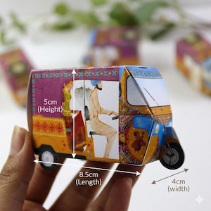 May include: A colorful paper model of an auto rickshaw, measuring 8.5cm long, 4cm wide, and 5cm high. The design features illustrations of people and intricate patterns in shades of yellow, blue, purple, and red. The model has small wheels and a detailed front.