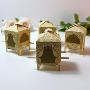 Puede incluir: Cuatro cajas decorativas doradas con forma de palanqu&iacute;n. Cada caja tiene un dise&ntilde;o ornamentado con un recorte en forma de ventana y clavijas de madera que se extienden desde los lados. Las cajas est&aacute;n dispuestas sobre una superficie blanca.