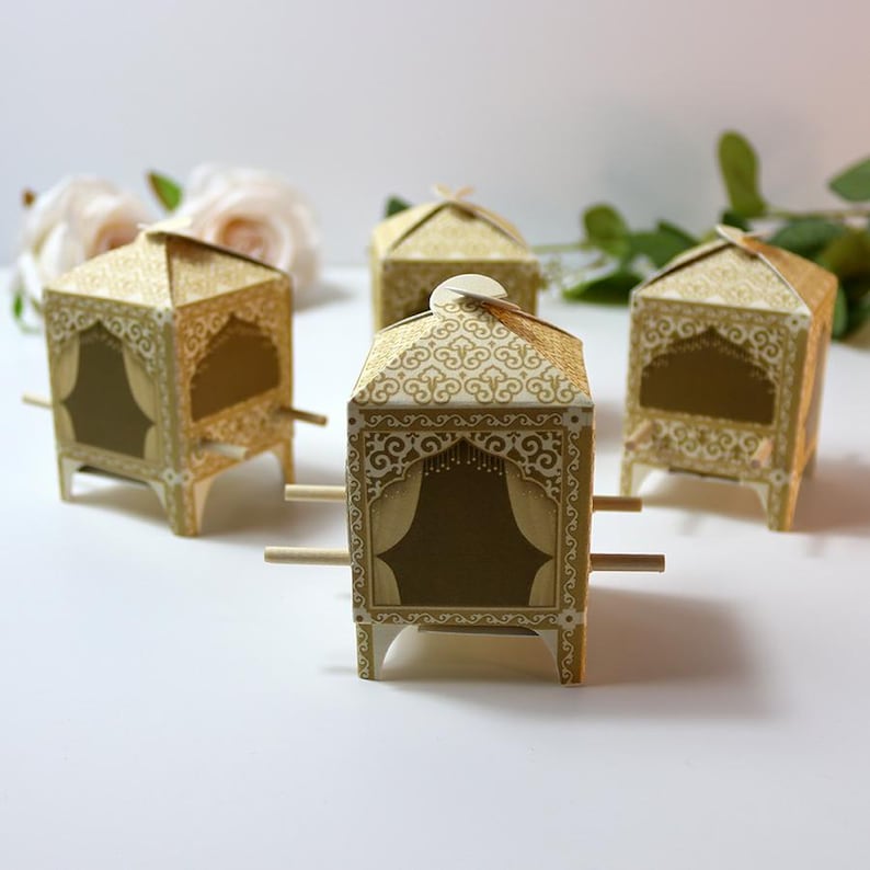 Puede incluir: Cuatro cajas decorativas doradas con forma de palanqu&iacute;n. Cada caja tiene un dise&ntilde;o ornamentado con un recorte en forma de ventana y clavijas de madera que se extienden desde los lados. Las cajas est&aacute;n dispuestas sobre una superficie blanca.