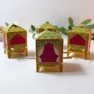Puede incluir: Cuatro estructuras decorativas de papel en miniatura amarillas y rojas con dise&ntilde;os ornamentados. Cada una tiene un techo puntiagudo y est&aacute; sostenida por peque&ntilde;os tacos de madera. Las estructuras est&aacute;n dispuestas sobre una superficie blanca, con vegetaci&oacute;n en el fondo.