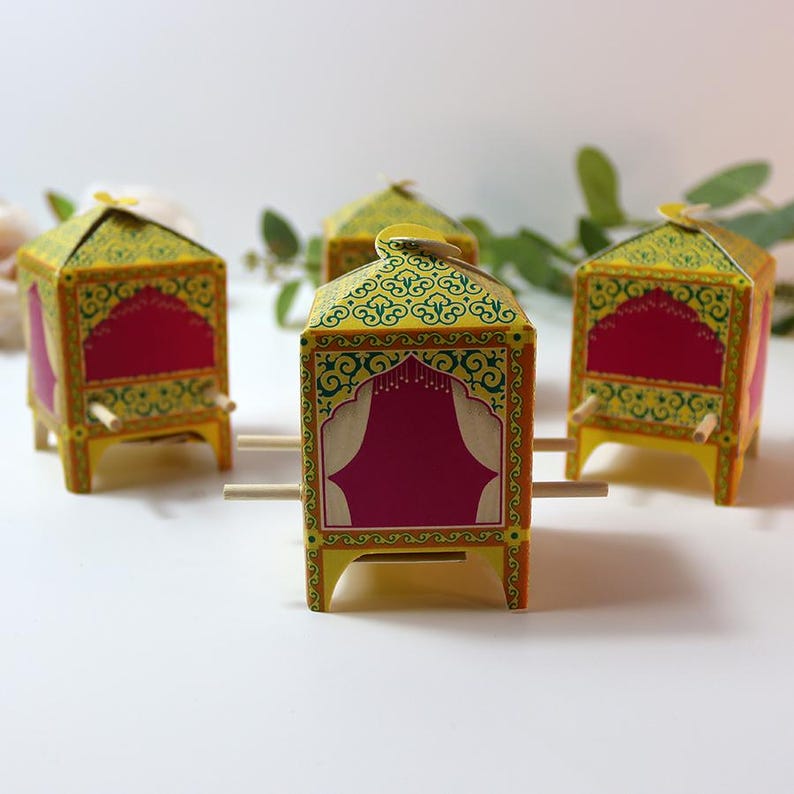 Puede incluir: Cuatro estructuras decorativas de papel en miniatura amarillas y rojas con dise&ntilde;os ornamentados. Cada una tiene un techo puntiagudo y est&aacute; sostenida por peque&ntilde;os tacos de madera. Las estructuras est&aacute;n dispuestas sobre una superficie blanca, con vegetaci&oacute;n en el fondo.