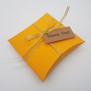 May include: A vibrant yellow gift box, secured with a gold ribbon and a small brown tag bearing the words "Thank You!". The box is square with curved edges, and the ribbon is neatly tied.