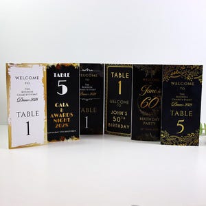 May include: A collection of six rectangular event signs featuring black, white, and gold colour schemes. The signs display various text including Table 1, Table 5, Gala & Awards Night 2028, John's 50th Birthday, and Jane's 60th Birthday Party.